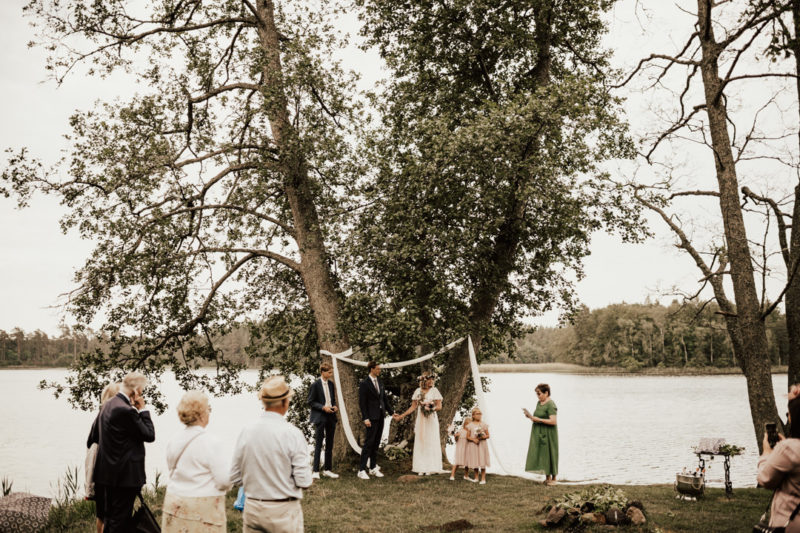 brudfölje och gäster vid utomhusvigsel vid sjö på midsommarafton