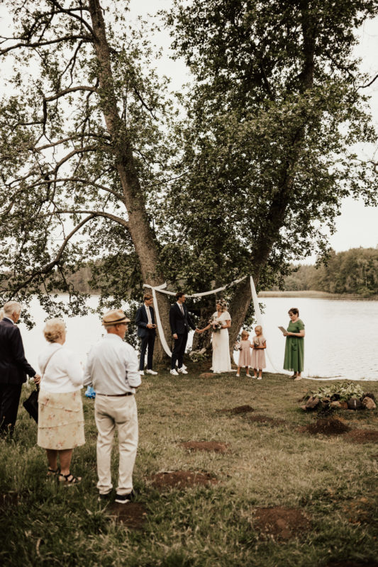brudfölje och gäster vid utomhusvigsel vid sjö på midsommarbröllop