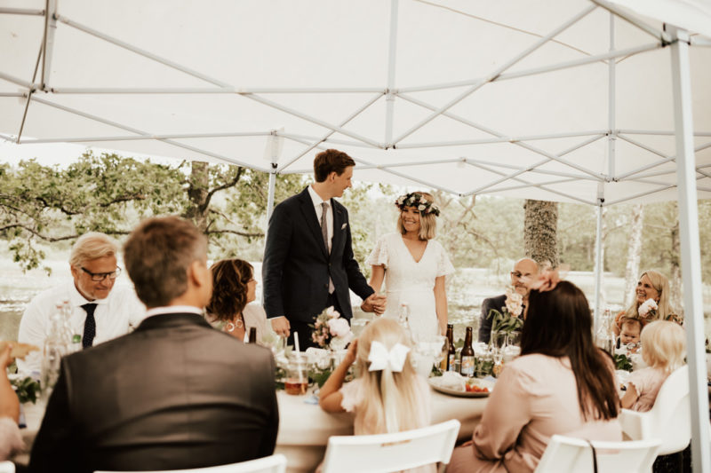 brudpar hälsar gäster välkomna på brunch utomhus på midsommarbröllop