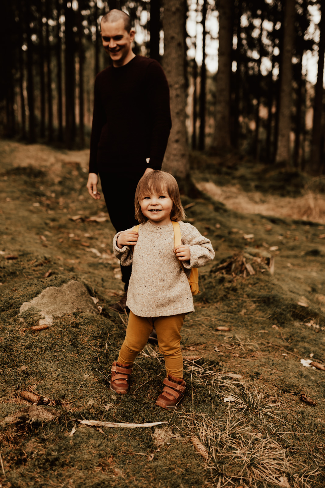 Barn med gula byxor och gul Fjällräven kånken står i skogen tillsammans med förälder