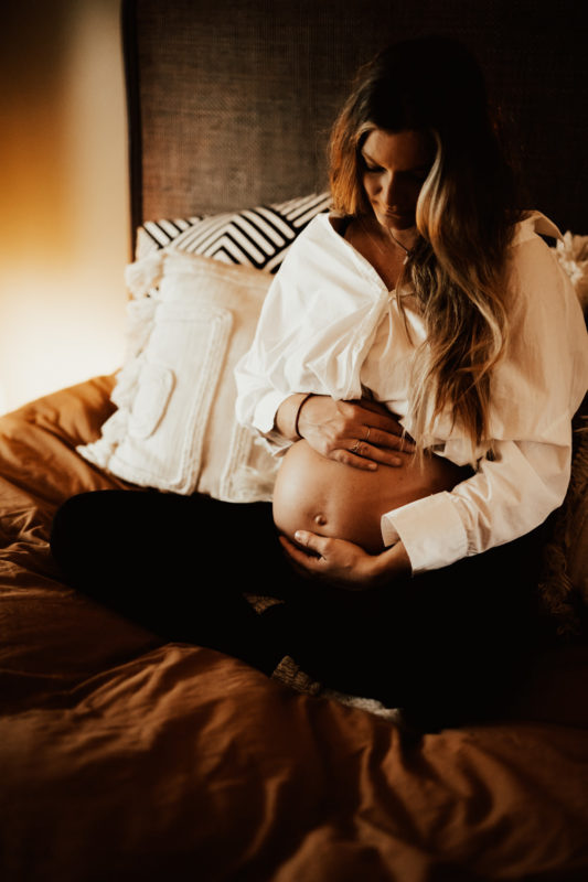 Gravid sitter i säng och håller om mage under gravidfotografering