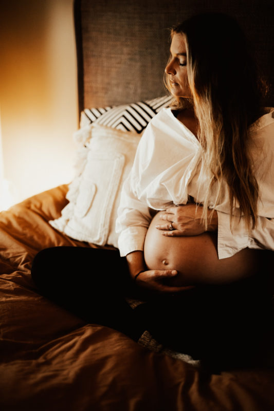 Gravid sitter i säng och håller om mage under gravidfotografering