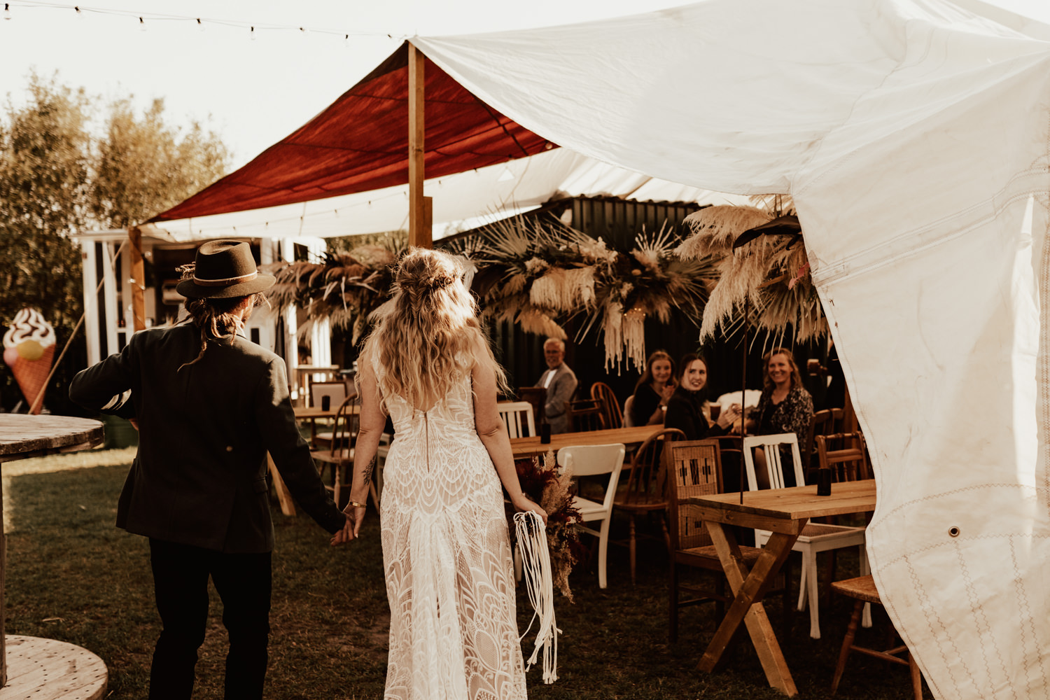 Wedding couple walks in to wedding venue at their outdoor festival wedding with sail stretch tent and hanging flower arrangements