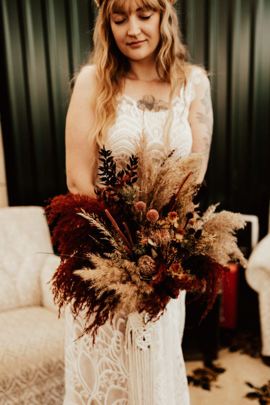 Boho bride standing in lace dress holding her bouquet made of dried flowers and pampas grass