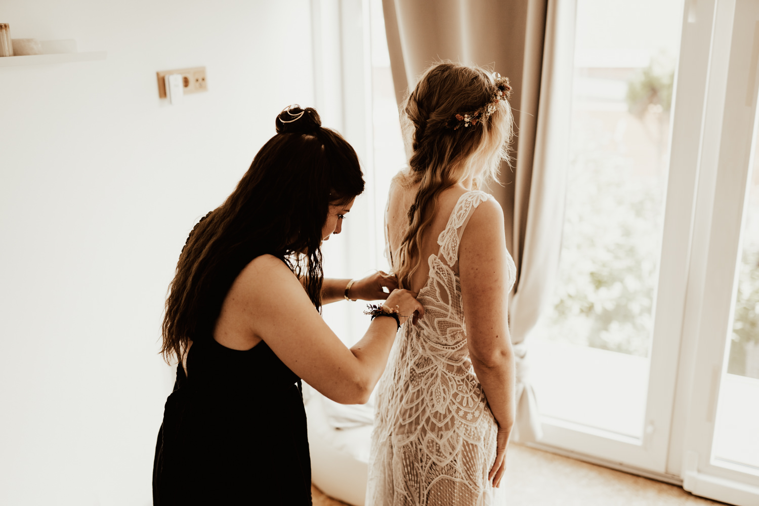 Bride standing faced to the window while bridesmaid buttons her lace boho dress before the wedding