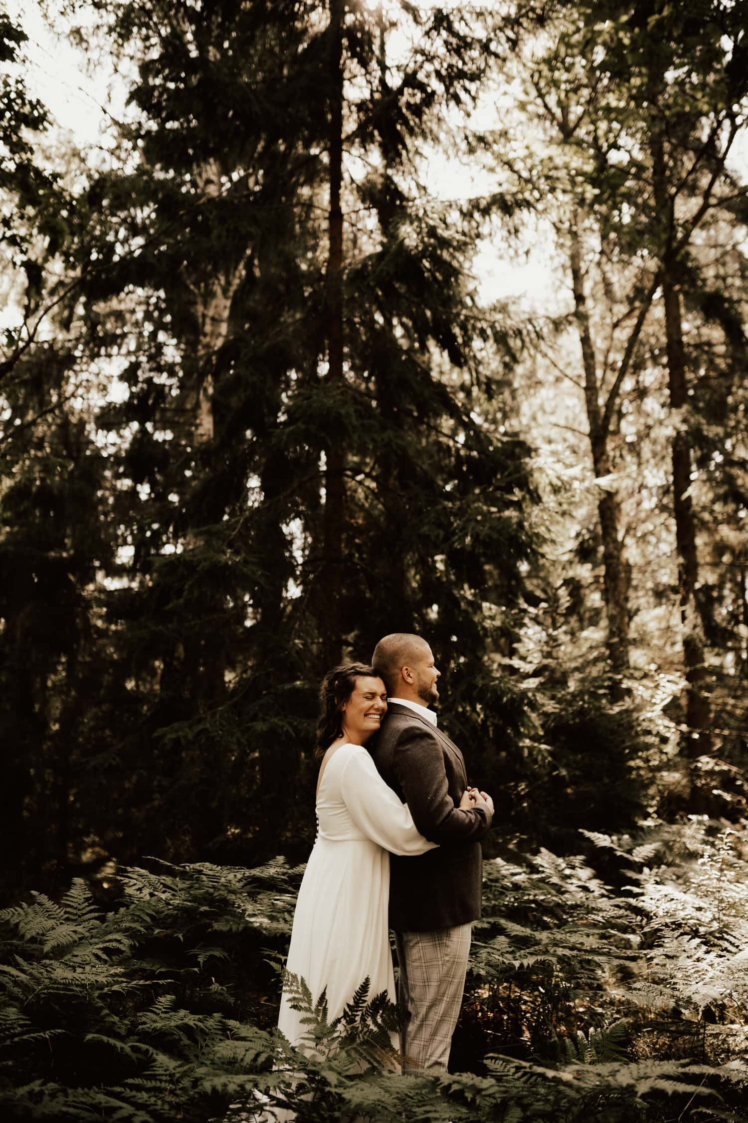 Brud kramar om brudgummen bakifrån medan de står i skog i Göteborg under deras bröllop