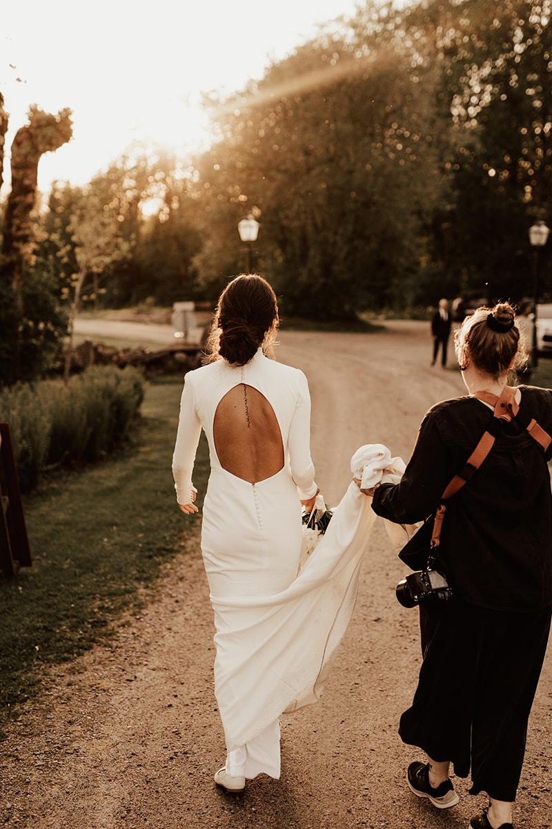 Bröllopsfotograf hjälper brud med brudklänningen medan de går mot kvällssolen