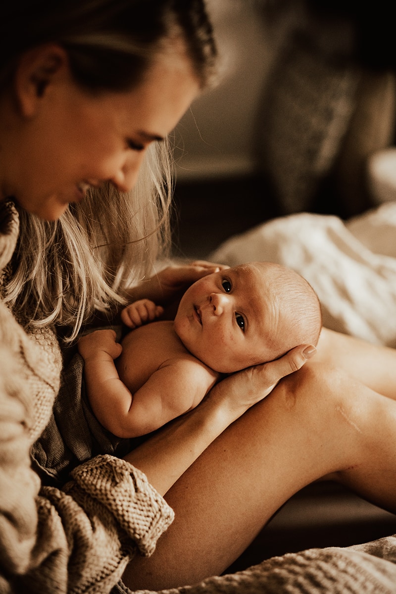 mamma sitter med bebis i knät och tittar på den under nyföddfotografering i hemmet