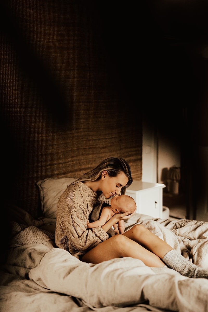 mamma sitter med beige stickad tröja i sin säng med nyfödd bebis i famnen och pussar på bebisen