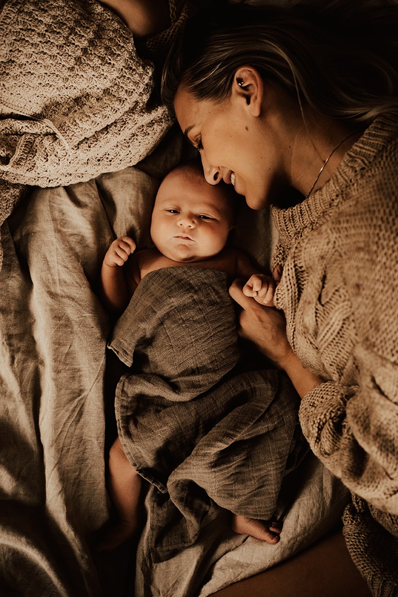 mamma ligger bredvid och ler mot sin nyfödda bebis som är insvept i brun linne filt