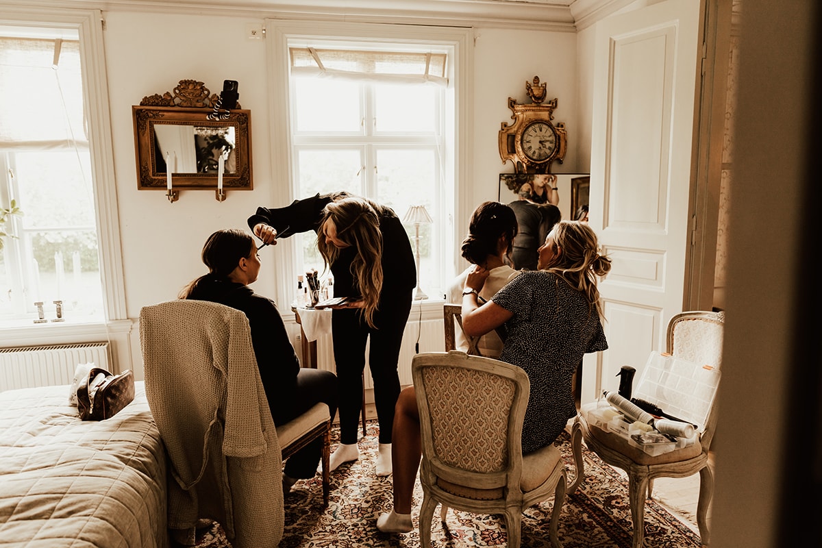 Brud och tärna sitter i stort rum och stylas av makeupartist och frisör innan bröllopsdagen på Uddetorp Säteri