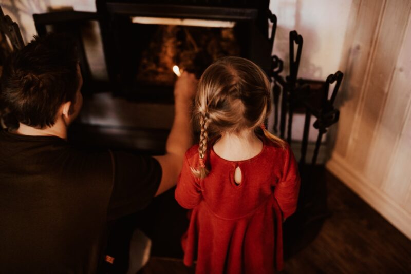 yngre dotter i röd klänning och flätor står bredvid pappa medan han tänder brasan under lifestylefotografering hemma.