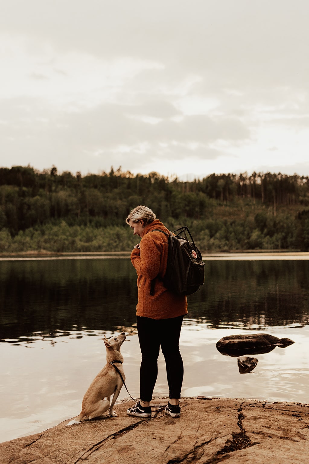 Kvinna i orange tröja och med grön ryggsäck står vid sjö och tittar ner mot sin lilla hund som tittar tillbaka.