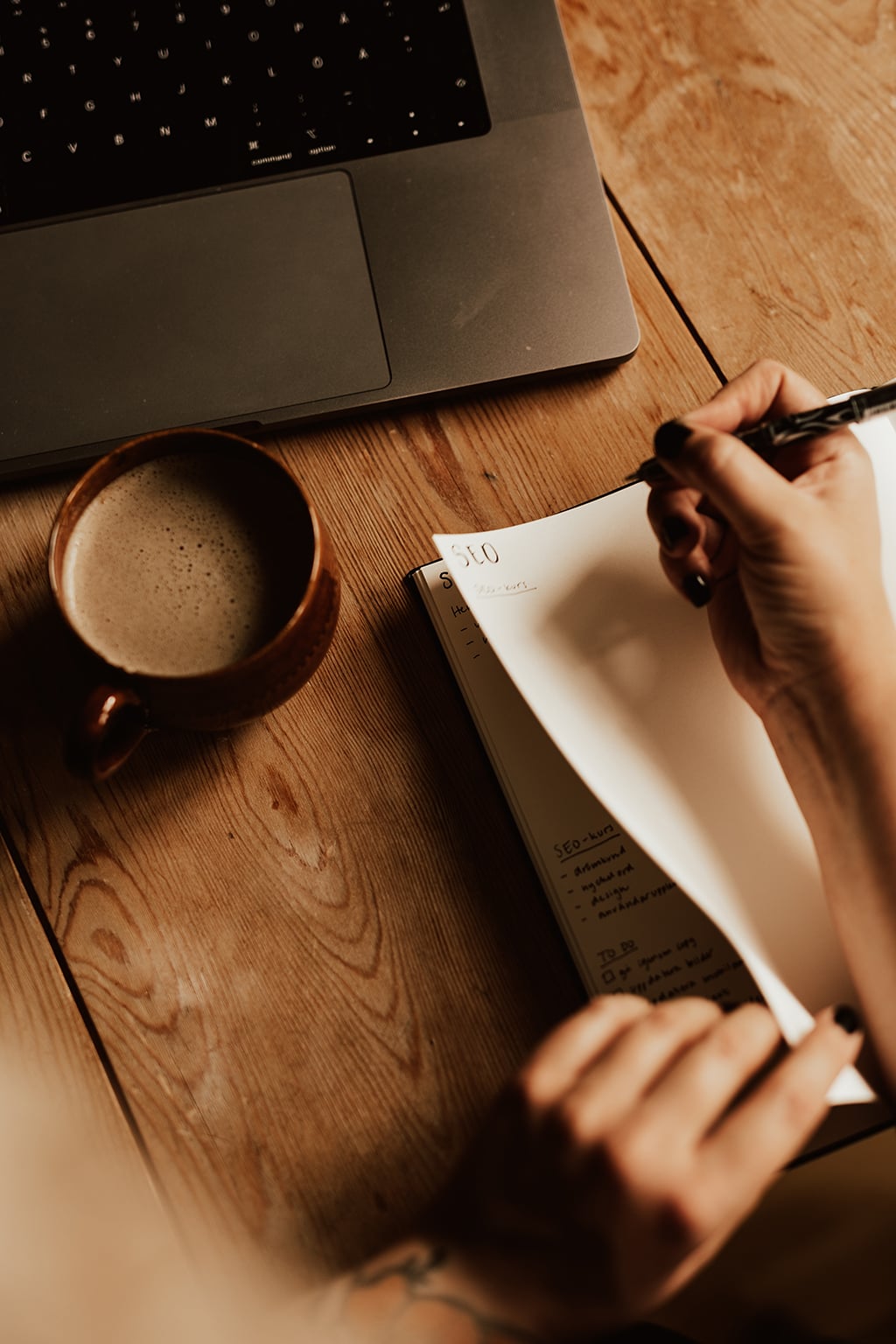 Person skriver SEO i sitt anteckningsblock med en kopp kaffe och dator bredvid.