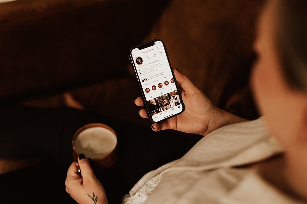 Fotograf sitter i brun fåtölj och håller i mobiltelefon och en kopp kaffe i andra handen och tittar ner mot mobiltelefonen.