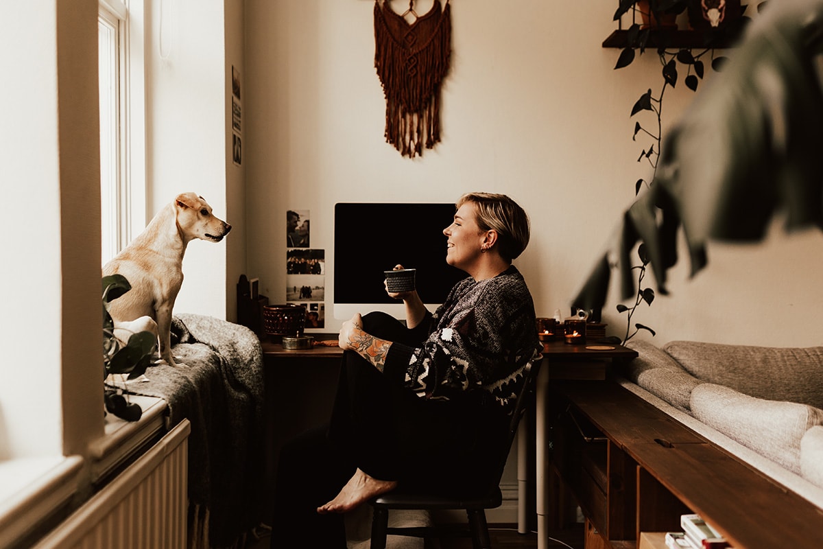 Kvinna sitter på skrivbordsstol med en kopp framför skrivbord, vänd mot en hund som sitter i fönstret. 