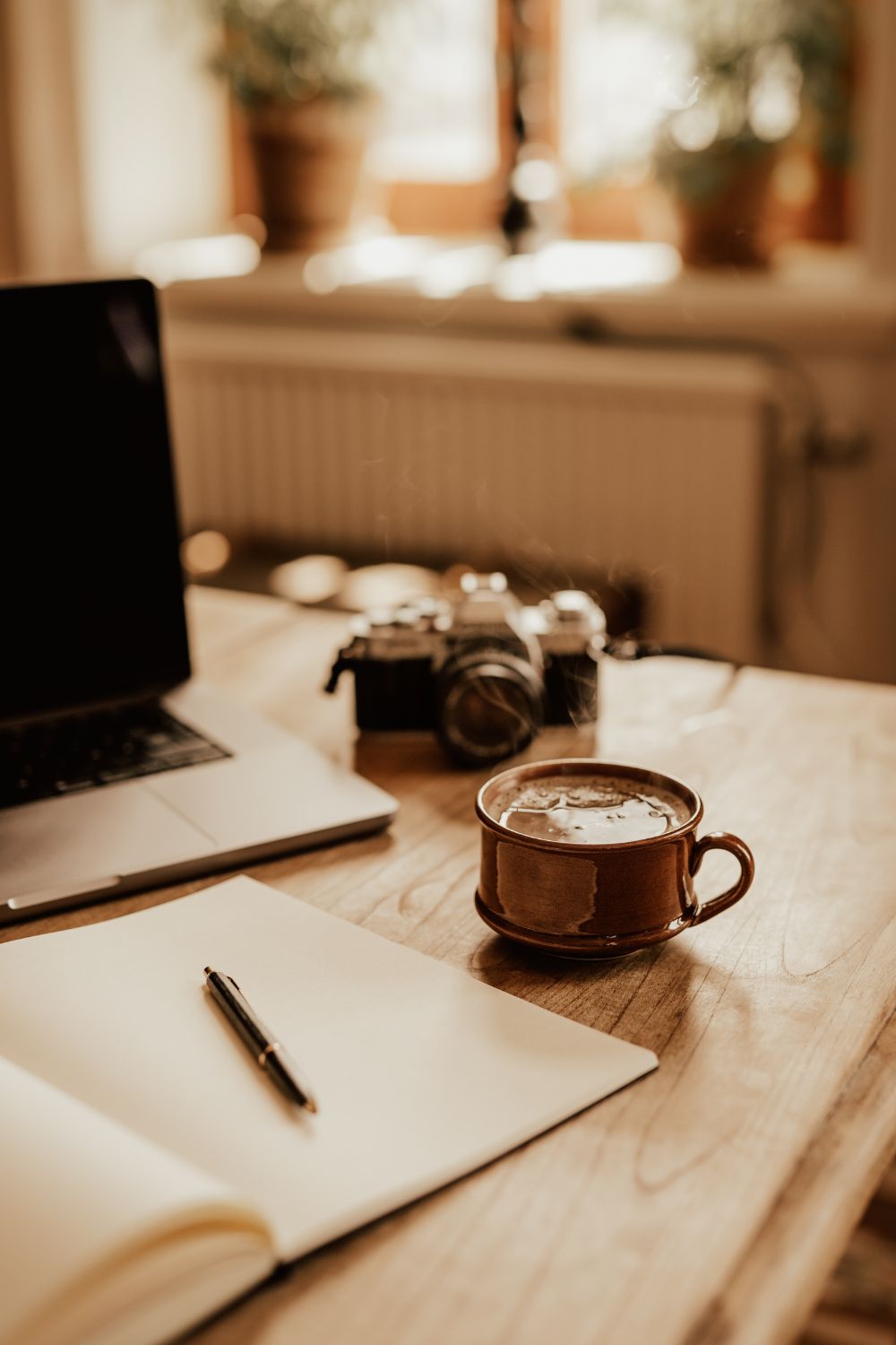 Kaffekopp, analog kamera, anteckningsbok och lap top på ett träbord.