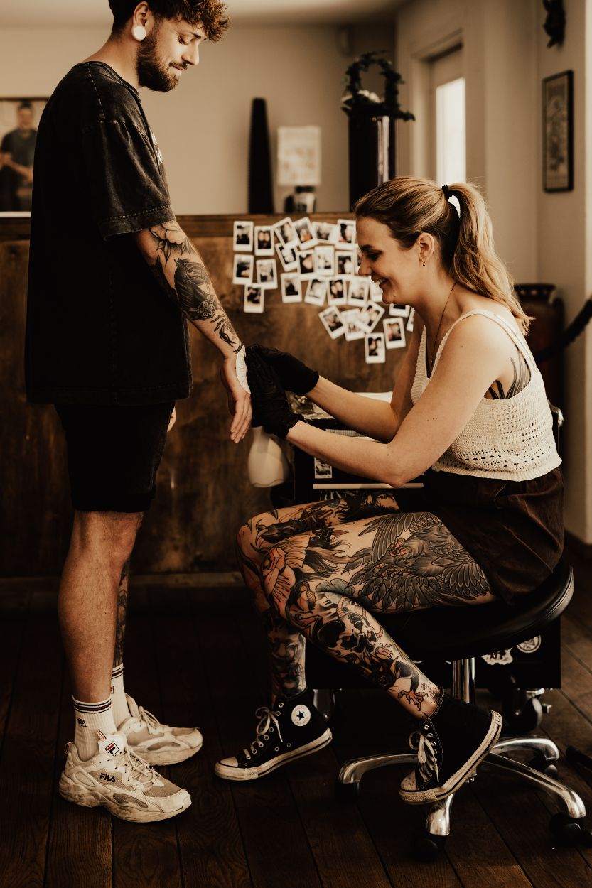 Kvinnlig tatuerare sitter på stol medan hon sätter på stencil på sin kunds hand i en tatueringsstudio.